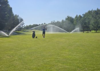 A golfer swings on a well-maintained green while sprinklers provide water on a sunny afternoon.